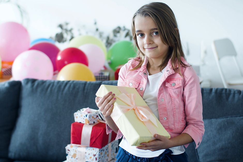 A young girl in a pink jacket enjoys her birthday celebration with gifts and colorful balloons indoors.